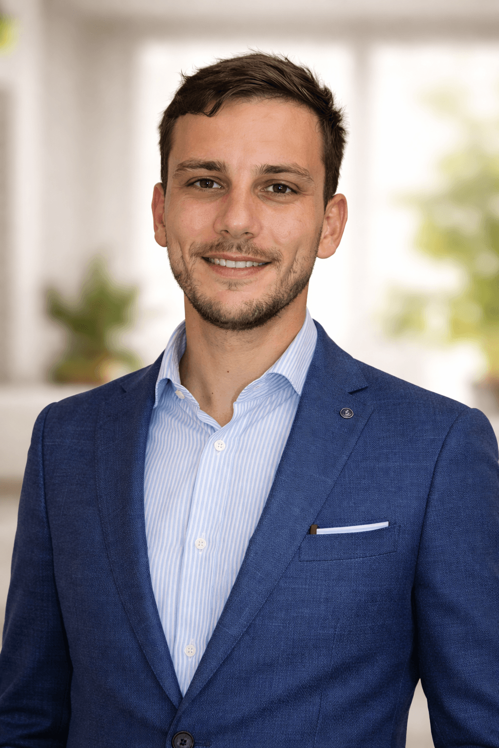 Smiling man with short brown hair wearing a blue blazer and striped dress shirt.
