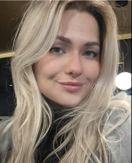 Young woman with long blonde hair and green eyes smiling softly in a grey jacket.